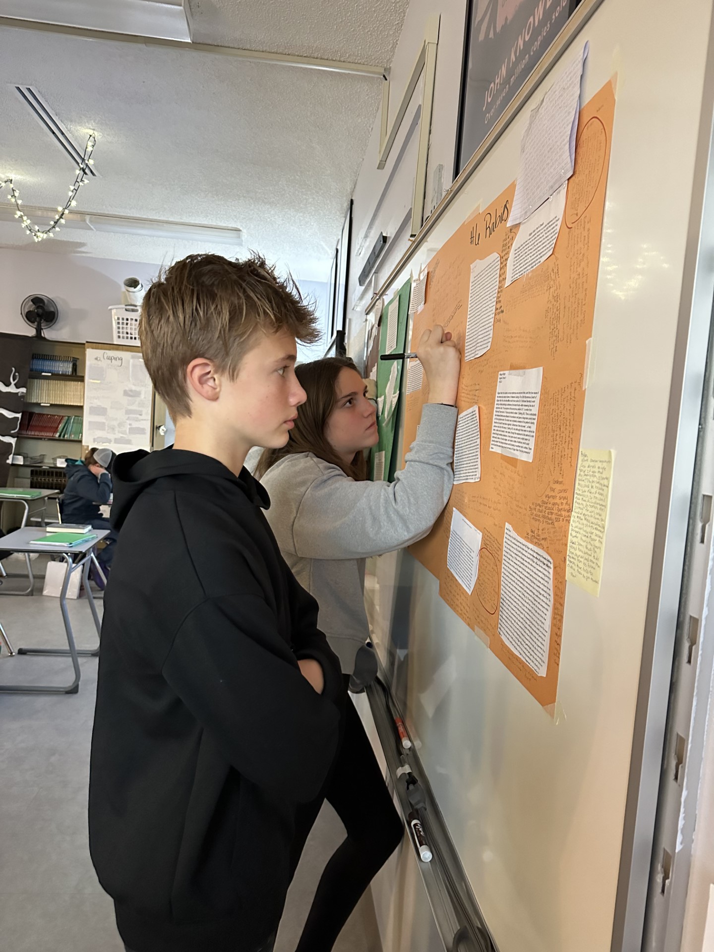Two Middle School English students stand by posters on wall. One is adding paragraph arguments to the poster, the other is reading existing paragraphs on the poster. 