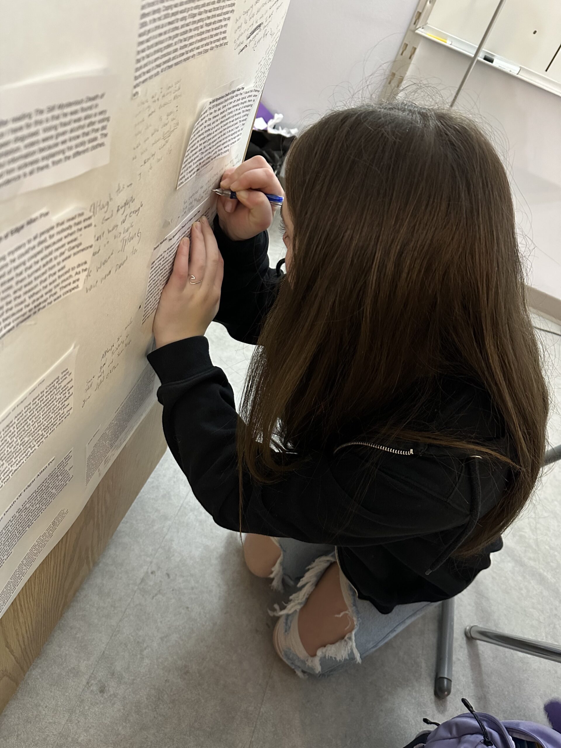 Middle School English student is kneeling by poster on a wall while writing paragraph arguments on the poster. 