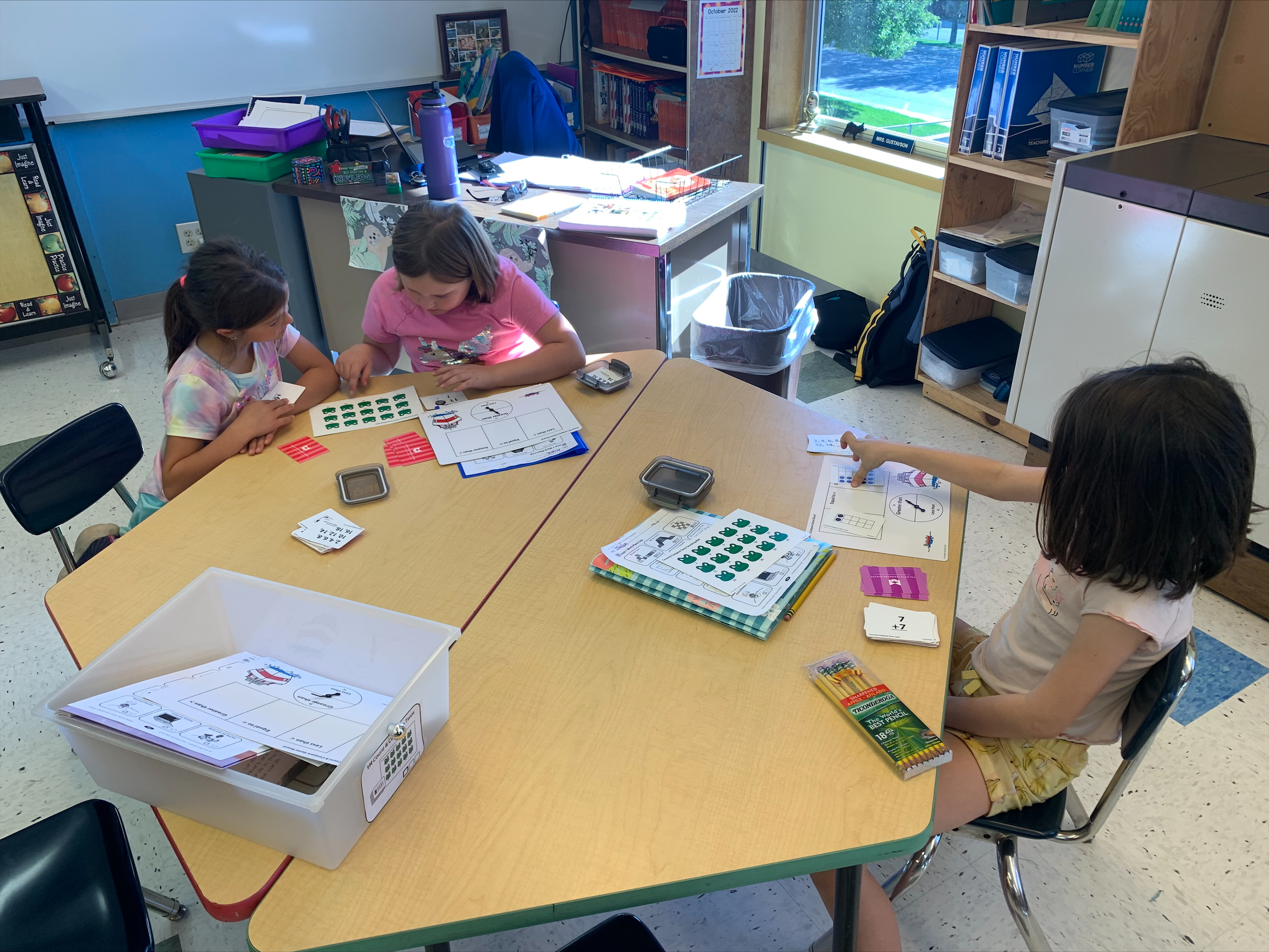 Two elementary students sit at one end of a table playing a math game. On the other side of the table is another student doing an individual math activity.