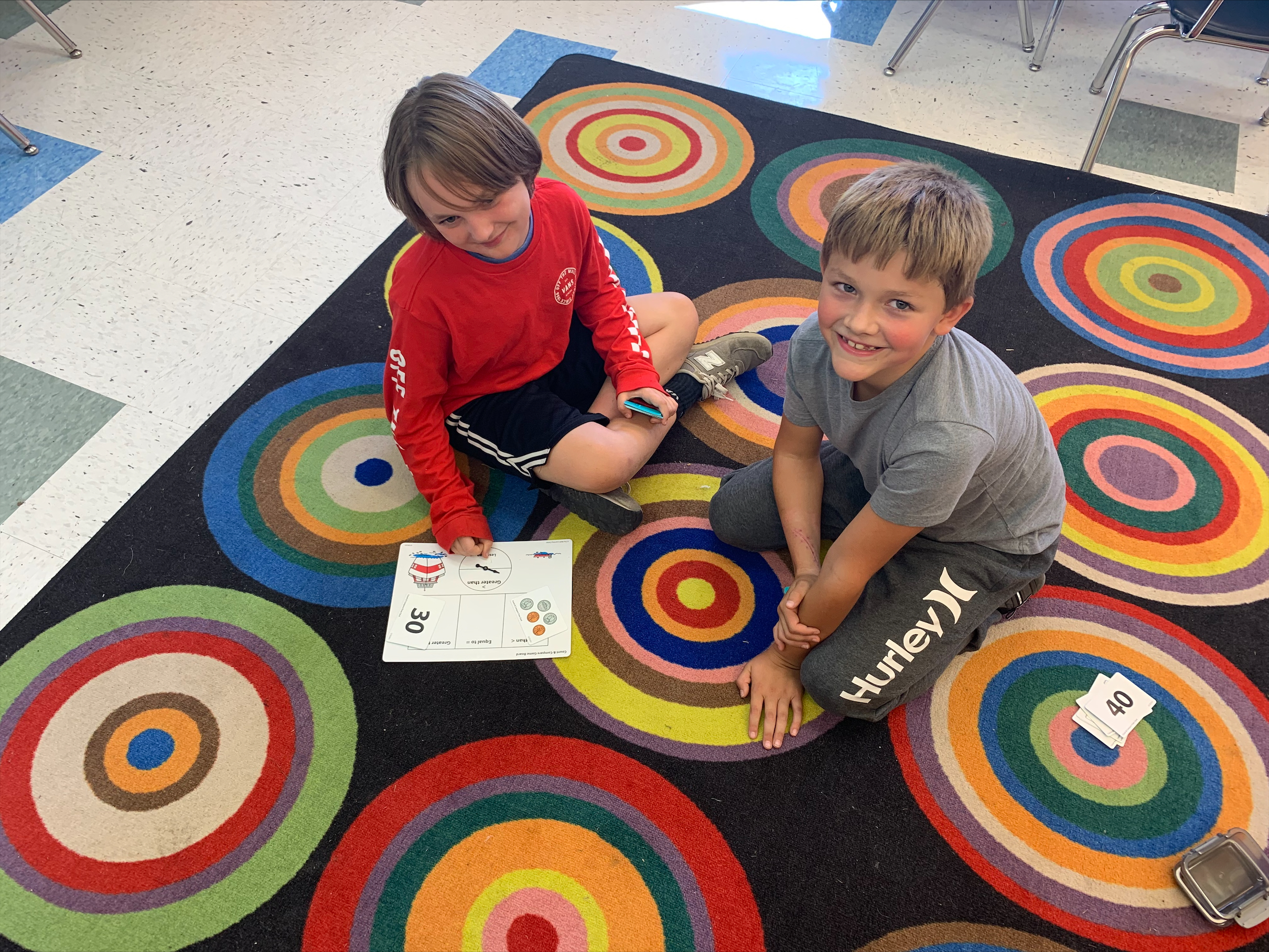 Two elementary students sit on a circle patterned mat playing math games.