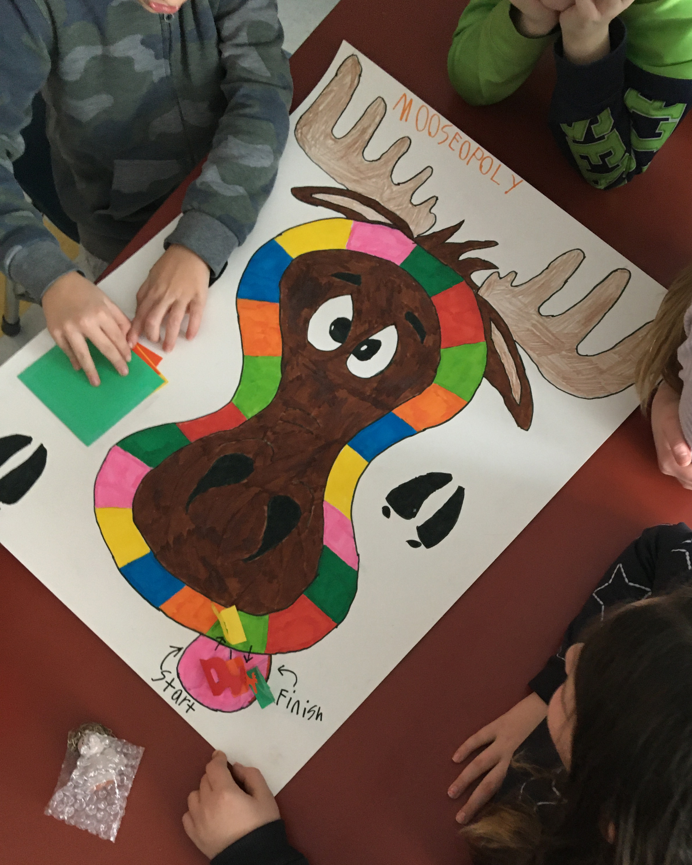 Students' hands shown as they play a student made board game in the shape of a moose.