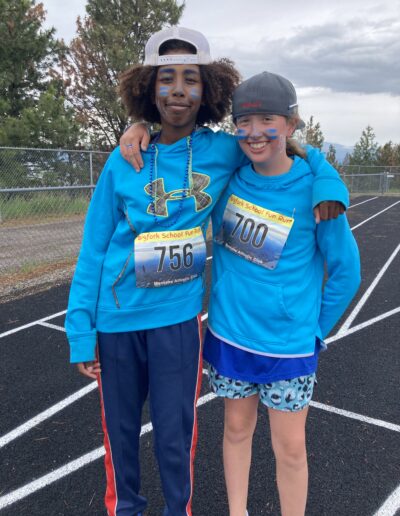 Two smiling students wearing blue and with blue face decorations stand with their arms around each others shoulders on the track during Fun Run.