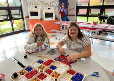 Two students sit at a table making bead jewelry at one of the booths at the fall carnival