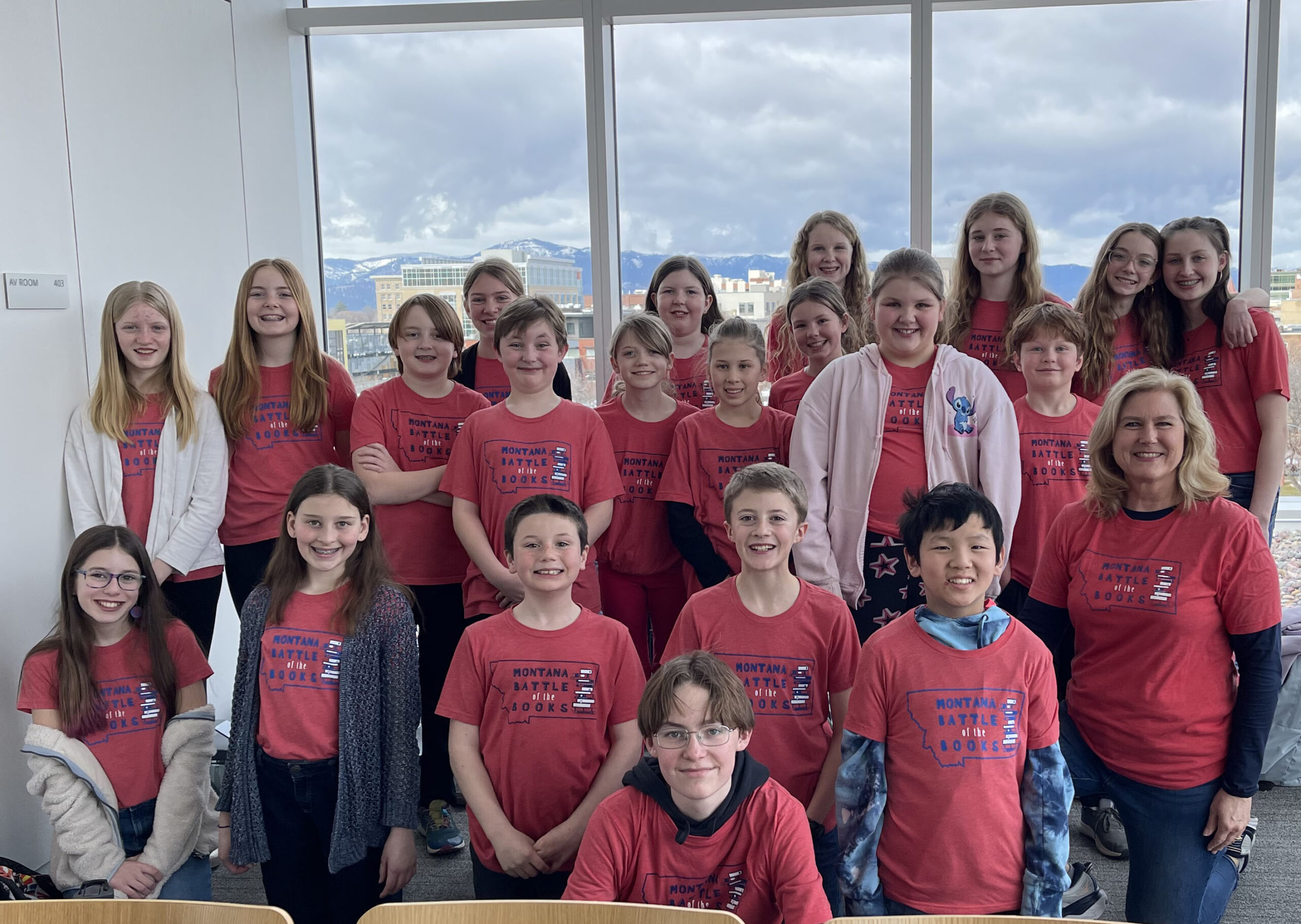 A large group of happy students participating in the 2024 Battle of the Books in Missoula. Behind them are big windows overlooking the city below.