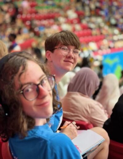 NHD students sit in the bleachers of a big stadium full of people. The students smile while looking back over their shoulders.