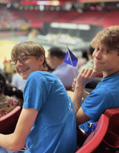 NHD students sit in the bleachers of a big stadium full of people. The students smile while looking back over their shoulders.