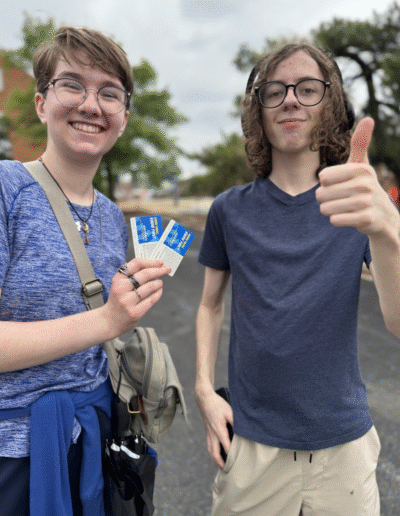 Two NHD students are standing on the tarmac with trees in the background. One is holding up two tickets and the other is giving a thumbs up.