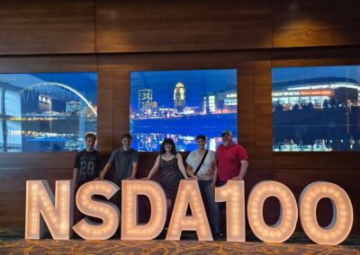 Speech and debate students and their coaches are standing behind a large display which says NSDA100. Behind them are images of a city skyline at night.
