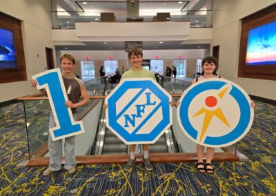 Three speech and debate students in the lobby of the competition venue holding up cut outs of the number one hundred.