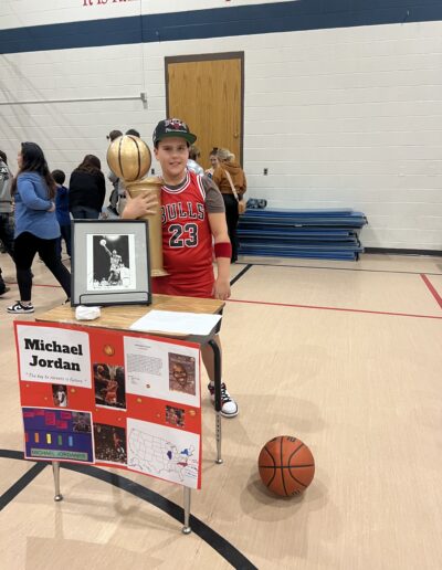 A student dressed as Michael Jordan and holding a trophy stands behind his Night of the Notables display.