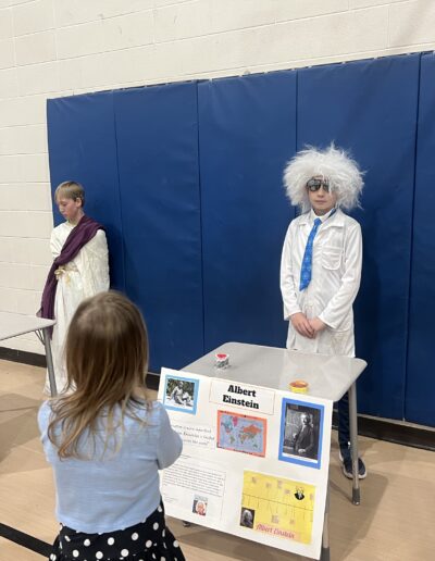 Three students dressed up as their Night of the Notables person stand behind their respective displays along the wall. A visitor is getting a closer look at the Albert Einstein display.