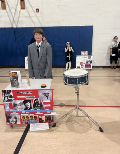A student dressed up as Ringo Starr stands behind his Night of the Notables display.