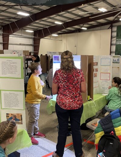 In a fairground building with high ceiling students are in conversation with science fair judges in front of their respective science fair display.