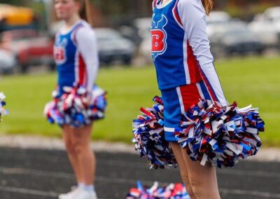 Two cheerleaders wearing cheer uniforms stand on a running track. Their arms are by their sides and they are holding poms in their hands.