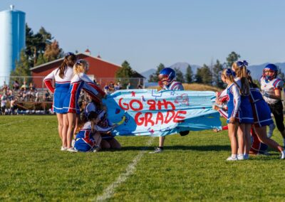 On a football field, a group of cheerleaders stand and sit on either side of a banner as they are holding it out between them. The banner says "Go 8th Grade" as two football players run toward the banner to be welcomed to the field.