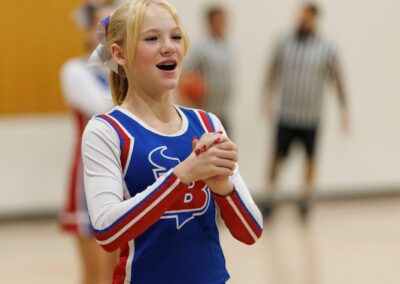 Two cheerleaders clasp their hands together as part of a cheer performed in a school gymnasium. Two referees are seen in the background.