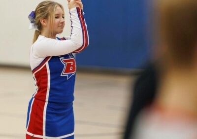 A cheerleader seen from the side, clasps her hands together in front of her face as a part of a cheer performance in a school gymnasium.