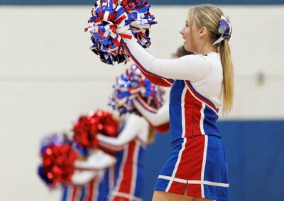 A row of cheer leaders perform a cheer in front of a gymnasium wall displaying a school logo.