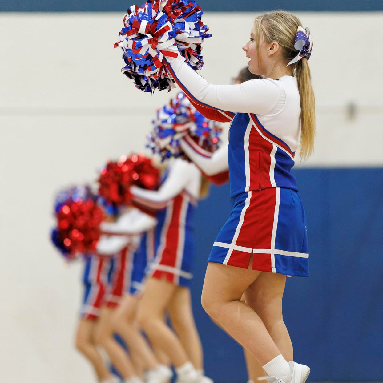 A row of cheer leaders perform a cheer in front of a gymnasium wall.